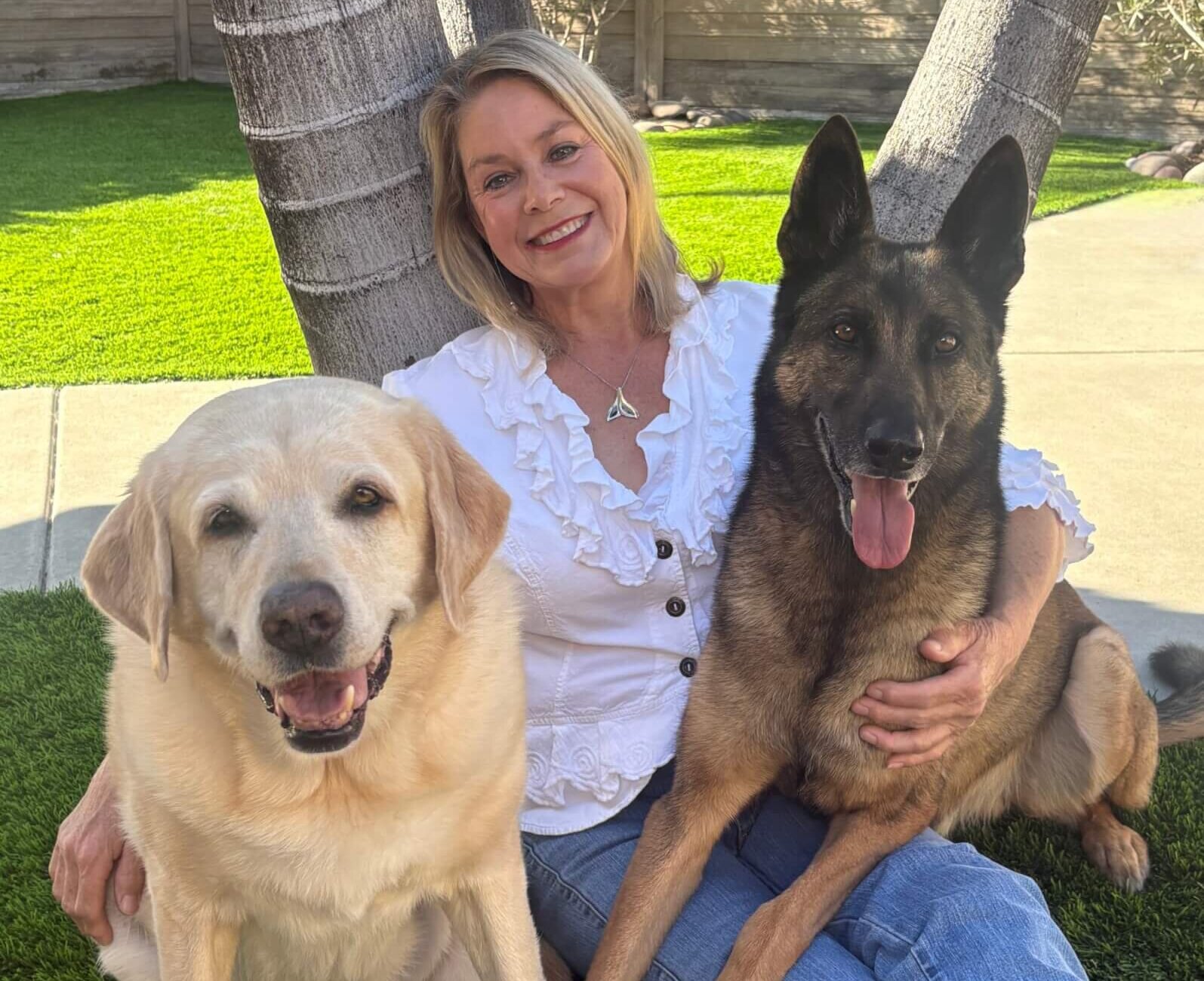 IMG_1397(1) A woman in a white blouse sits on grass, leaning against a tree, smiling with a yellow Labrador and a German Shepherd by her side.